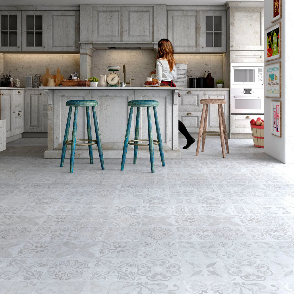 A wide interior kitchen scene with pale gray patterned flooring, white cabinets, two teal bar stools at the island, and a woman standing near the counter.