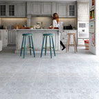 A wide interior kitchen scene with pale gray patterned flooring, white cabinets, two teal bar stools at the island, and a woman standing near the counter.
