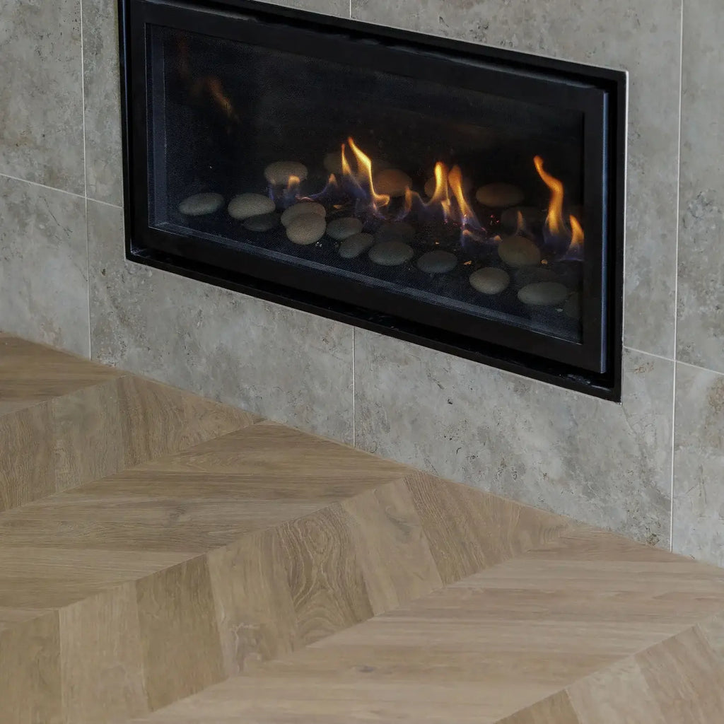 A modern linear gas fireplace with a black frame, set into a light beige stone tiled wall above a light oak herringbone wood floor. The fireplace contains smooth dark stones and active orange and yellow flames.