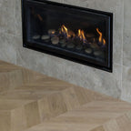 A modern linear gas fireplace with a black frame, set into a light beige stone tiled wall above a light oak herringbone wood floor. The fireplace contains smooth dark stones and active orange and yellow flames.