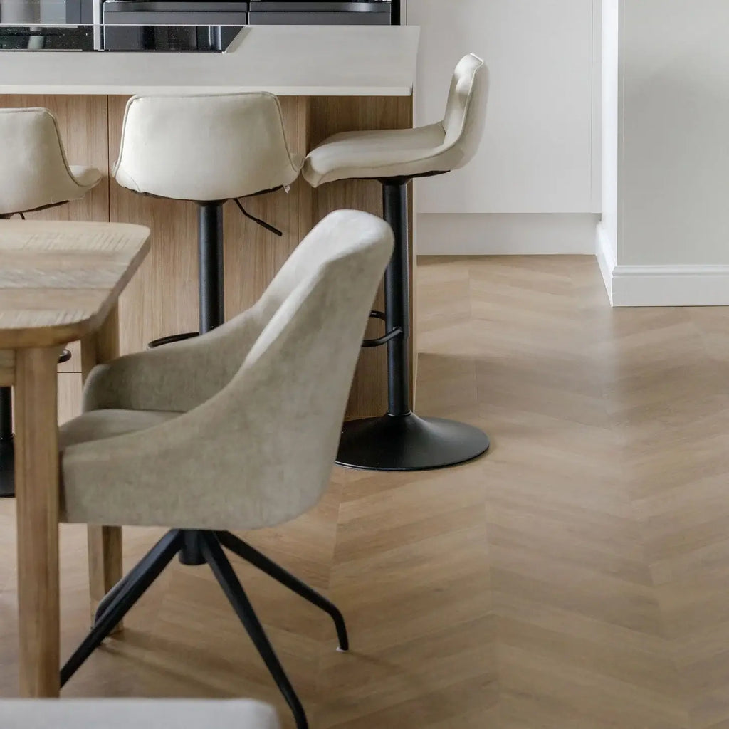 A modern dining room scene featuring a light beige velvet swivel chair in the foreground, with two matching high-back bar stools and a wooden island in the background, set on a light herringbone wood floor.