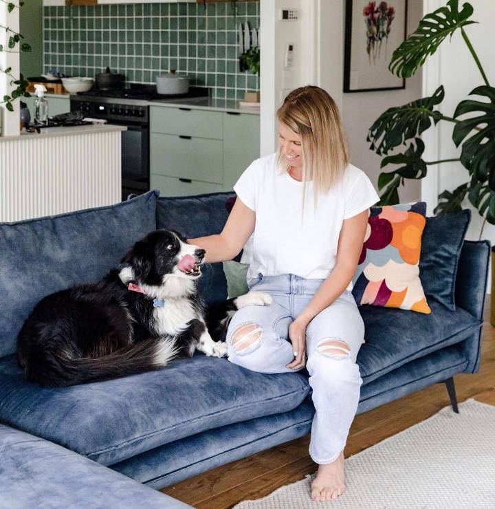 A woman smiles and pats her border collie on a blue velvet sofa in a warm, contemporary living room with beautiful timber flooring, lush indoor plants, and a stylish open-plan kitchen in the background.
