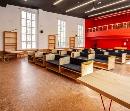 A spacious contemporary commercial interior featuring warm cork flooring that adds natural texture and acoustic comfort underfoot, complemented by bold red feature walls, timber furnishings, and large windows that flood the space with natural light.