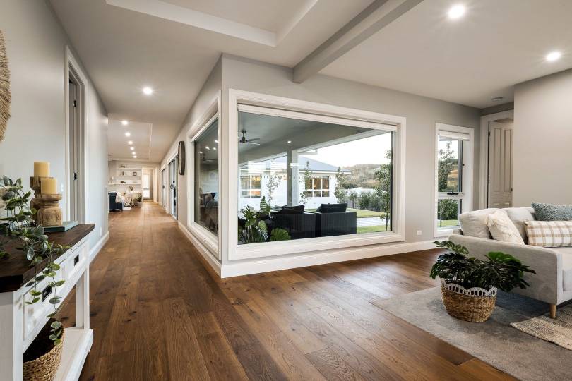 A spacious, light-filled Hampton-style home interior showcasing rich dark oak hardwood timber flooring that runs the full length of a grand open-plan living area and hallway, complemented by white-framed picture windows, neutral furnishings, and leafy indoor plants.