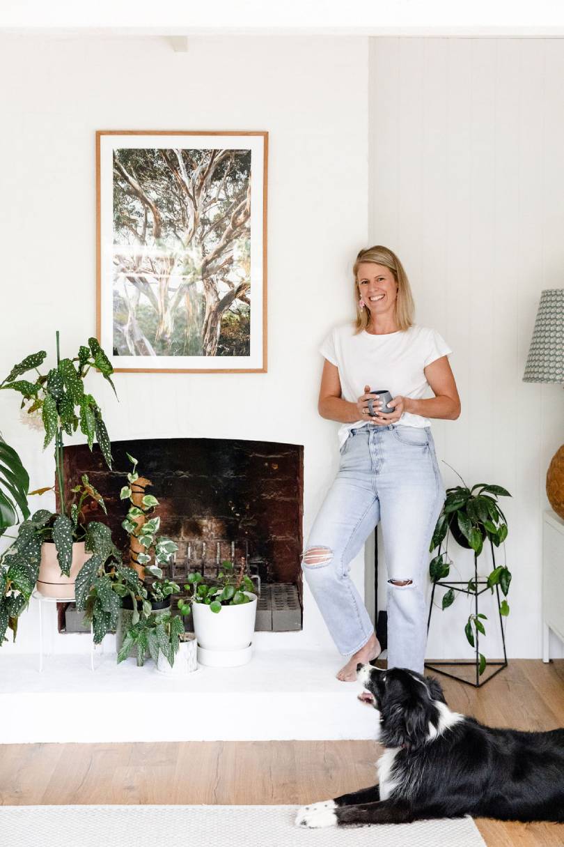 A smiling woman enjoys a coffee beside a charming fireplace filled with lush indoor plants, her border collie resting contentedly on a rug over warm natural timber flooring, creating a relaxed and inviting Australian home interior full of personality and life.
