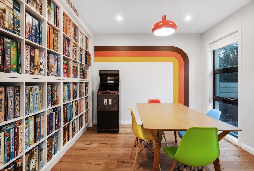 A fun and vibrant retro-inspired games room with warm natural hardwood timber flooring, featuring a floor-to-ceiling board game library, a vintage arcade machine, colourful Eames-style chairs, and a bold 1970s rainbow mural that brings personality and playfulness to the space.