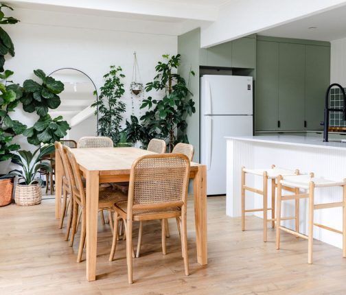 A relaxed, nature-inspired open-plan dining and kitchen space with light blonde laminate flooring, styled with a rustic timber dining table, woven rattan chairs, olive green cabinetry, and an abundance of lush indoor plants for a fresh, organic feel.
