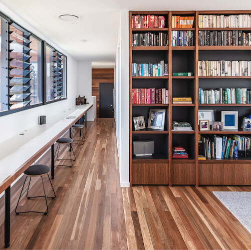 A stylish home study and library featuring rich, dark Australian hardwood timber flooring with stunning natural grain variation, paired with a floor-to-ceiling walnut bookshelf, a sleek white wall-mounted desk, and black wire stools for a sophisticated, functional workspace.