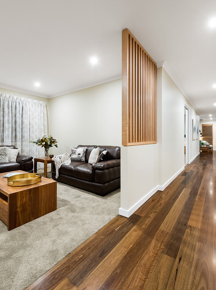 A modern Australian home interior where rich dark hardwood timber flooring flows through a long hallway and transitions seamlessly into a carpeted lounge room, cleverly divided by a striking timber batten screen that adds warmth, texture, and architectural interest.
