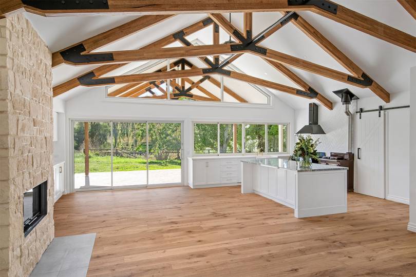 A breathtaking open-plan rural home interior featuring warm natural hardwood timber flooring beneath soaring cathedral ceilings with exposed timber trusses and black steel joinery, complemented by a stone fireplace, white kitchen cabinetry, and expansive glass doors opening to a lush garden.