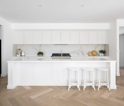 An elegant all-white Hamptons-style kitchen with a marble stone splashback and brass tapware, anchored by stunning light oak herringbone parquetry timber flooring that adds warmth, texture, and timeless sophistication to the classic monochromatic design.