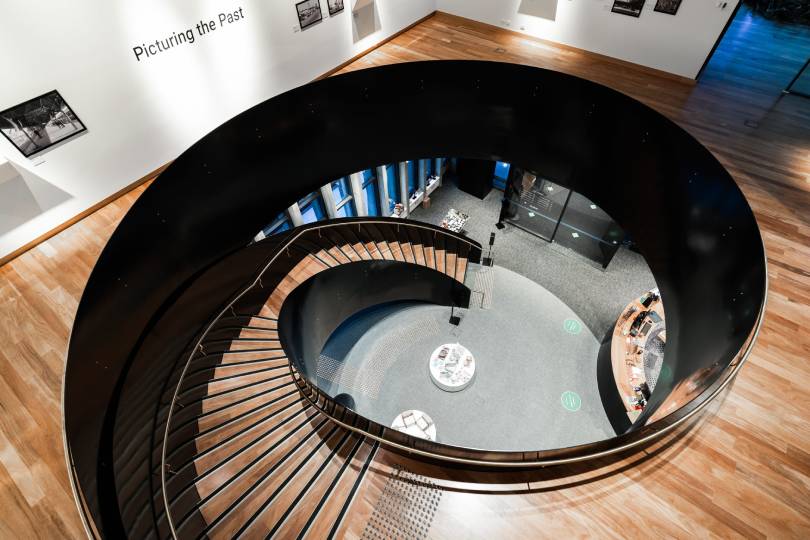 A dramatic overhead view of a sweeping spiral staircase with warm timber flooring and stair treads contrasting beautifully against bold black steel framework, set within a grand public building interior that showcases the versatility and elegance of hardwood timber floors in commercial spaces.