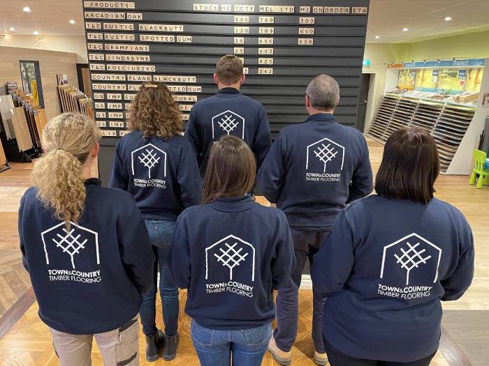 The friendly Town & Country Timber Flooring team standing together in their navy branded uniforms inside their timber flooring showroom, surrounded by an extensive range of hardwood flooring samples and a full product stock board on display.